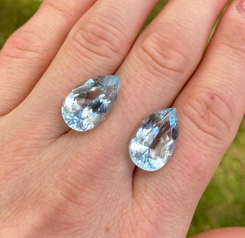 May include: Two pear-shaped, faceted aquamarine gemstones. The gemstones are a light blue color and have a high degree of clarity. The stones are displayed on a light skin tone background.