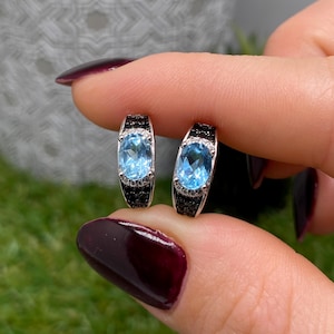May include: A pair of silver earrings featuring oval, light blue gemstones. The earrings are detailed with black stones. The earrings are held between fingers against a blurred green background.