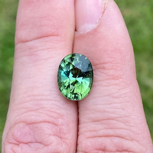 Puede incluir: Una gema ovalada y facetada con un tono verde vibrante, que exhibe un juego de luz y color. Las facetas de la gema crean un efecto brillante, mostrando su claridad y brillo. La gema se sostiene en una mano, con un fondo verde borroso.
