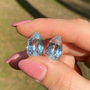 May include: Two pear-shaped, light blue gemstones. The gems are held between fingers with pink nail polish. The gems have facets that reflect light, creating a sparkling effect. The background is blurred, suggesting an outdoor setting.