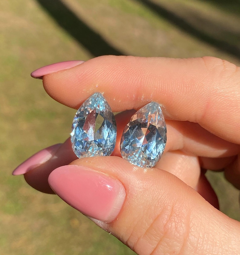 May include: Two pear-shaped, light blue gemstones. The gems are held between fingers with pink nail polish. The gems have facets that reflect light, creating a sparkling effect. The background is blurred, suggesting an outdoor setting.