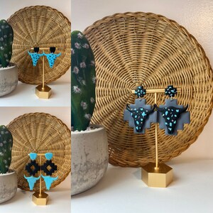 Puede incluir: Un par de pendientes con un diseño de calavera de vaca negra y turquesa. Los pendientes se muestran en un soporte dorado sobre un fondo tejido.