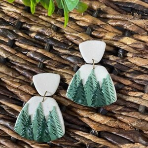 Puede incluir: Un par de pendientes hechos a mano con una parte superior blanca y una parte inferior con temática de bosque. La parte inferior presenta un diseño de bosque verde. Los pendientes se muestran sobre una superficie marrón tejida.