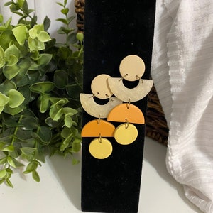 Puede incluir: Pendientes colgantes geométricos en tonos crema, naranja y amarillo. Los pendientes presentan una combinación de formas circulares y semicirculares, conectadas por pequeños aros dorados. Se exhiben sobre una superficie de terciopelo negro.