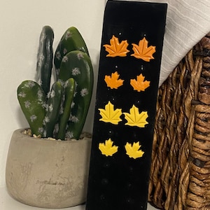 Puede incluir: Exhibición de cuatro pares de pendientes con forma de hoja en tonos naranja y amarillo. Los pendientes están sobre un expositor de terciopelo negro. Un cactus en maceta y una cesta tejida se encuentran en el fondo.