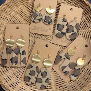 Puede incluir: Cuatro pares de pendientes hechos a mano con un diseño de estampado de leopardo y detalles dorados. Los pendientes se muestran en cartulina marrón con el texto "CLAYxGEORGIANA" impreso en cada tarjeta.
