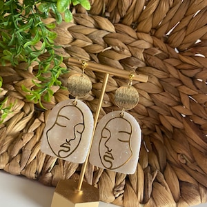 Puede incluir: Un par de pendientes dorados y blancos con un diseño de rostro minimalista. Los pendientes están en un soporte dorado sobre un fondo tejido.