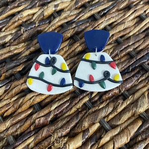 Puede incluir: Un par de pendientes festivos con luces navideñas. Cada pendiente tiene una base blanca adornada con luces de colores rojo, amarillo, verde y azul, con cableado negro. Cada pendiente tiene un triángulo azul oscuro en la parte superior. Para orejas perforadas.