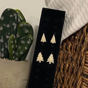 Puede incluir: Cuatro pares de pendientes con forma de árbol de Navidad sobre un expositor negro. Los pendientes son de color claro y brillante. Un cactus en maceta y una cesta de mimbre están en el fondo.