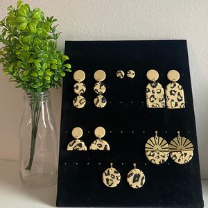 Puede incluir: Una muestra de varios pendientes de leopardo dorados y negros. Los pendientes se muestran en una tabla negra con fondo blanco. Los pendientes están hechos de arcilla polimérica y son ligeros y cómodos de llevar.