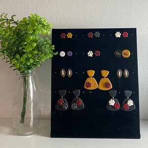 Puede incluir: Un tablero de exhibición negro con varios pendientes de arcilla de colores. Los pendientes tienen forma de huellas de patas, balones de fútbol y formas abstractas. Los pendientes están dispuestos en filas en el tablero.