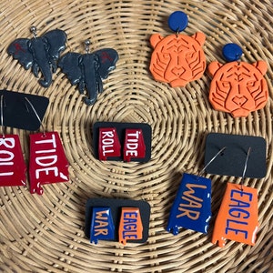 Puede incluir: Un conjunto de 10 pares de pendientes con varios logotipos y mascotas de equipos universitarios. Los pendientes están hechos de arcilla polimérica y vienen en una variedad de colores, incluyendo rojo, azul, naranja y negro. Los pendientes tienen forma del estado de Alabama, una cara de tigre, un águila y un rollo de papel. Los pendientes son perfectos para mostrar tu espíritu de equipo.