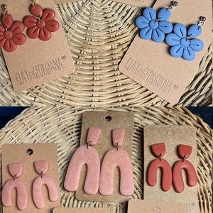 Puede incluir: Surtido de pendientes hechos a mano de arcilla polimérica en varios colores y formas, incluyendo flores, arcos y diseños abstractos. Los pendientes se muestran en cartulina marrón con el texto "CLAY by GEORGIANA" en cada tarjeta.