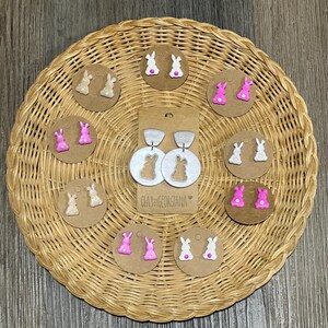 Puede incluir: Surtido de pendientes de conejito de arcilla polimérica hechos a mano en rosa y blanco. Los pendientes se muestran en una superficie de mimbre con el texto "Clay by Georgiana" en una tarjeta.