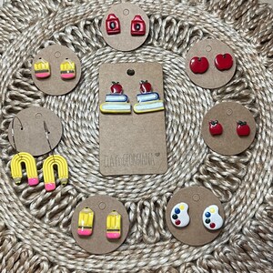 Puede incluir: Una colección de pendientes de arcilla polimérica hechos a mano sobre cartulina marrón. Los diseños incluyen manzanas rojas, lápices, libros con manzanas, paletas de pintura y escuelas rojas. Los pendientes están dispuestos sobre una superficie tejida beige.