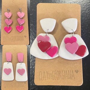 Puede incluir: Pendientes de arcilla polimérica hechos a mano en varios diseños. Un par presenta tres corazones rosas apilados, otro tiene una base blanca con un corazón rosa, y un tercer par tiene una base blanca con corazones rosas y rojos. Todos están en tarjetas marrones.