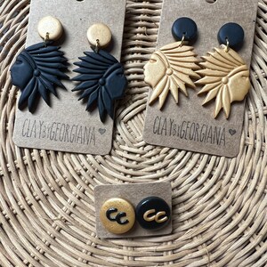 Puede incluir: Tres pares de pendientes hechos a mano de arcilla polimérica. Un par presenta un diseño de tocado nativo americano en negro y dorado. Otro par presenta un diseño de tocado nativo americano dorado. El tercer par presenta un diseño "CC" en negro y dorado.