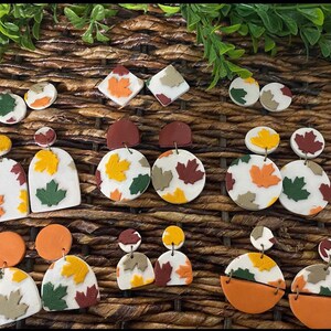 Puede incluir: Una colección de pendientes hechos a mano de arcilla polimérica en varias formas y tamaños. Los pendientes presentan diseños de hojas de otoño en tonos naranja, amarillo, verde y marrón, sobre fondos blancos y de color sólido. Los pendientes se exhiben sobre una superficie tejida.