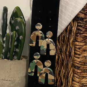 Puede incluir: Un par de pendientes hechos a mano con un patrón de camuflaje. Los pendientes tienen forma de arco y tienen un pequeño acento redondo en la parte superior.