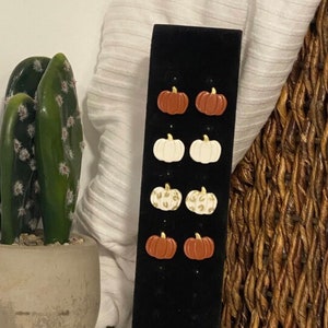 Puede incluir: Pendientes con forma de calabaza en marrón, blanco y estampado de leopardo. Se exhiben en un soporte de terciopelo negro, perfectos para accesorios de temática otoñal.