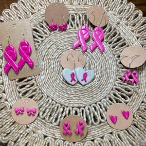 Puede incluir: Una colección de pendientes hechos a mano con un diseño de lazo rosa brillante. Los estilos incluyen tachuelas de lazo, aros de cinta y pendientes colgantes de cinta. Los pendientes se exhiben en pequeñas tarjetas redondas de cartón marrón.