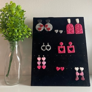 Puede incluir: Una exhibición de pendientes hechos a mano en varios estilos. Los pendientes presentan formas y patrones de corazón, incluyendo rayas y colores lisos. La paleta de colores incluye tonos de rosa, rojo, negro y blanco. Los pendientes se exhiben en una tabla de terciopelo negro.