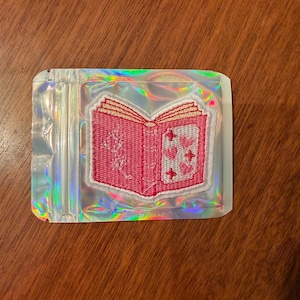 May include: A pink embroidered patch in the shape of an open book, with white trim and details. The book's pages are red, and the right page features hearts and a cross design. The patch is inside a clear, iridescent plastic bag.