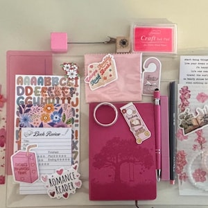 May include: A collection of pink and white stationery items, including a notebook, pens, stickers, and a stamp pad. The items are arranged on a light background, creating a visually appealing display. The theme appears to be related to reading and journaling.