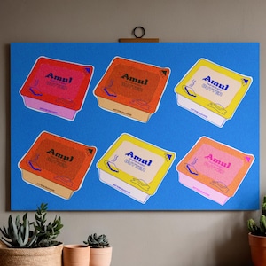 Puede incluir: Un lienzo azul muestra seis cajas de mantequilla Amul coloridas. Cada caja presenta el logotipo y el texto de Amul, con variaciones de color que incluyen rojo, naranja y amarillo. La obra de arte está montada en una pared con plantas y un cuadro enmarcado en el fondo.