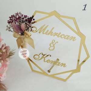May include: Gold geometric acrylic sign with the names "Mihrican & Kenan" in a cursive font. A small bouquet of dried flowers is attached to the sign.