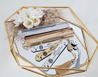 May include: A gold-colored tray with a mirrored surface holds three bookmarks with Islamic calligraphy and tassels. The bookmarks are in a decorative box with a white and gold design. The tray is decorated with white flowers and dried grass.