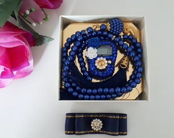 May include: A blue prayer bead set with a digital counter and a tassel. The counter has a white flower design and the words "COUNTER" and "RESET" are visible. The set is packaged in a white box with a gold interior.