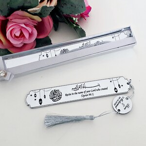 May include: A silver bookmark with a tassel and a quote from the Quran in Arabic and English. The bookmark is in a silver box with a white ribbon. The quote reads "Recite in the name of your Lord who created (Quran 96:1)".