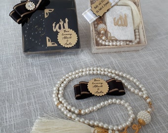 May include: A white prayer bead necklace with a white tassel, a black and gold bow, and a small white box with gold lettering that says "Dars Ceremony Ahmet & Aisha". The box is in a white gift box with a white tassel and a small white card that says "Baky-eid 2021 De Inés & Ayman".