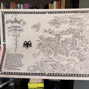 May include: A detailed black and white map of the Calradian Empire, featuring intricate geographical details, cities, and text. The map includes the title "THE DOMINIONS CALRADIAN EMPIRE" and decorative borders. The map is held by a person.