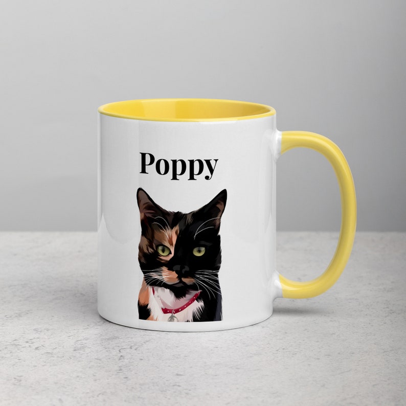 May include: A white ceramic mug with a yellow rim and handle. The mug features a colorful illustration of a cat with the name "Poppy" printed in black text.