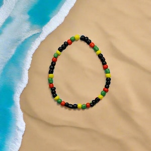 Może przedstawiać: Bransoletka z koralików z czarnymi, czerwonymi, żółtymi i zielonymi koralikami. Bransoletka leży na piaszczystej powierzchni, z niebieską falą oceanu w tle. Koraliki ułożone są w okrąg.