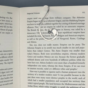 Könnte beinhalten: Ein silberfarbener Metall-Lesezeichen in einem einzigartigen, geschwungenen Design, das auf den Seiten eines Buches liegt. Die Form des Lesezeichens erinnert an ein stilisiertes Fragezeichen oder ein fließendes Band und verleiht dem Lesen eine elegante Note.