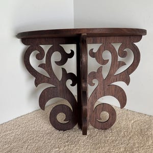 Może przedstawiać: Drewniana półka narożna w ciemnym kolorze brązu z misternymi detalami w postaci zwijanych wzorów. Półka wykonana jest z litego drewna i ma naturalne wykończenie.