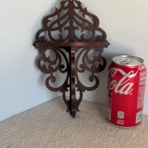 Puede incluir: Una estantería de madera marrón oscuro con intrincados recortes en forma de árbol estilizado. La estantería está montada en una pared y tiene una pequeña repisa en la parte superior. Una lata de Coca-Cola está sentada en la estantería.