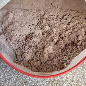 May include: A close-up of a brown powder in a red plastic container. The powder is likely a food product, such as cocoa powder or protein powder.