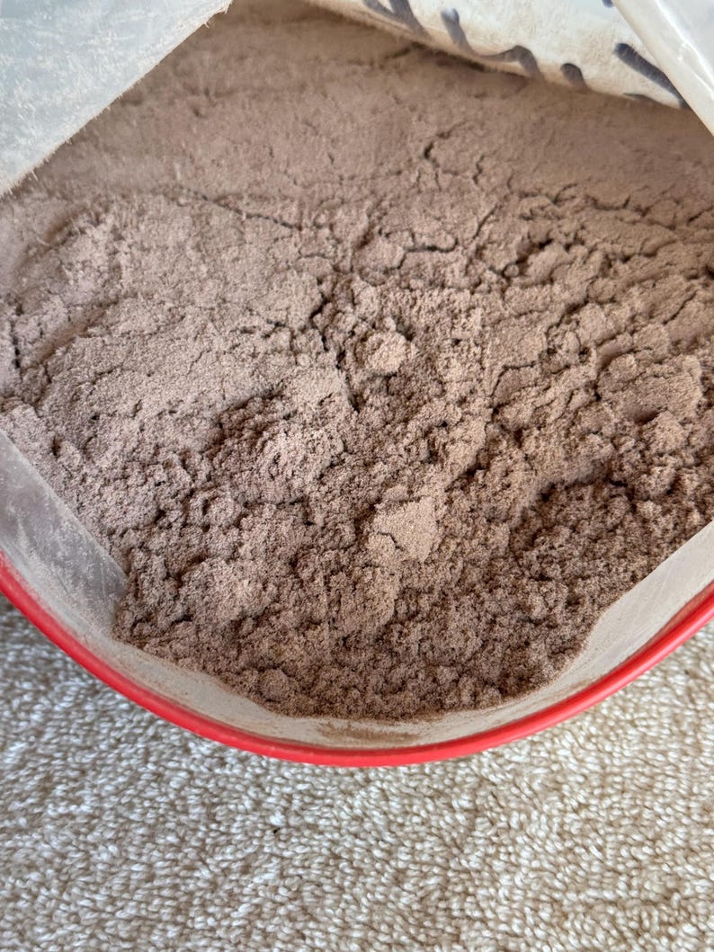 May include: A close-up of a brown powder in a red plastic container. The powder is likely a food product, such as cocoa powder or protein powder.