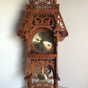 Puede incluir: Un reloj de madera con tallas intrincadas y un péndulo. La esfera del reloj tiene números romanos y un fondo de color dorado.