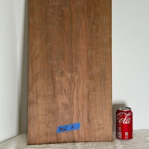 May include: A rectangular piece of light brown wood veneer with a visible natural grain pattern. A small blue piece of tape is attached to the lower section with the handwritten text 3/16" A. A red soda can is positioned next to the wood board.