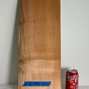 May include: A rectangular piece of light brown wood with a natural grain pattern. A blue piece of painter's tape is attached to the bottom left corner with the handwritten text A-1/4-8 3/4 x 20 1/2. A red Coca-Cola soda can is placed next to the wood.