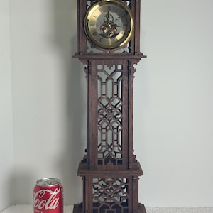 May include: A dark brown wooden grandfather clock with a gold-colored clock face. The clock has intricate carved details and a decorative top. A can of Coca-Cola is next to the clock for scale.