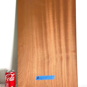 May include: A rectangular piece of reddish-brown wood with a visible grain pattern. A blue label is attached to the surface with the text A - 1/16" x 4 3/4" x 24". A red soda can is positioned next to the wood for scale, showing the dimensions.
