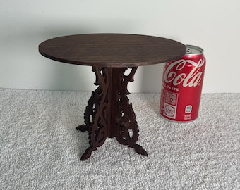 May include: A small, dark brown wooden table with a round top and intricately carved legs. The table is approximately 12 inches in diameter. A red Coca-Cola can is next to the table for size comparison.