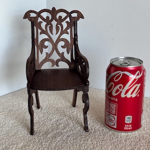 May include: A miniature wooden chair with an ornate, open-work back and curved legs. The chair is dark brown and stands next to a red Coca-Cola can, 12 fl oz (355 mL). The chair is a decorative item.