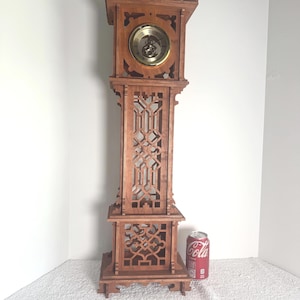 May include: A tall, brown wooden grandfather clock featuring intricate laser-cut geometric lattice patterns on the front panels and a circular gold-toned clock face. A standard red soda can is placed next to the base for scale reference.